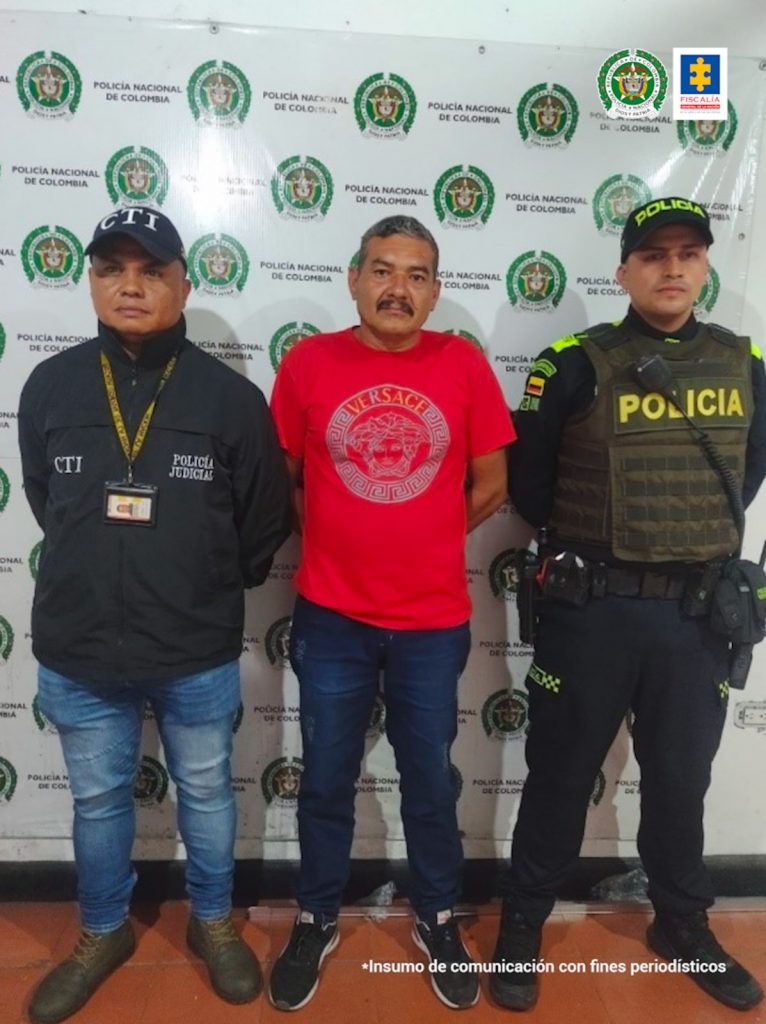 En la foto se aprecia de pie y con los brazos atrás a Norbey García Valencia. Viste camiseta de manga corta de color rojo, con estampado circular gris; jean azul oscuro y tenis negros con blanco. Al lado derecho de él hay un servidor del CTI de la Fiscalía y al lado izquierdo un uniformado de la Policía Nacional. Detrás de ellos hay un pendón institucional de la Policía.