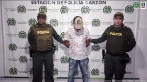 en la imagen se ve una persona detenida bajo custodia de dos integrantes de la Policía Nacional. Detrás suyo un backing institucional.