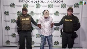  en la imagen se ve una persona detenida bajo custodia de dos integrantes de la Policía Nacional. Detrás suyo un backing institucional. 