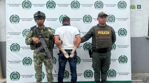  El imputado se encuentra de espaldas y tiene un buso blanco y jean y las manos esposadas y tiene las manos vendadas, se encuentra en custodia por un uniformado de la Policía y un uniformado del Ejército Nacional. Detrás se observa un backing de la Policía.