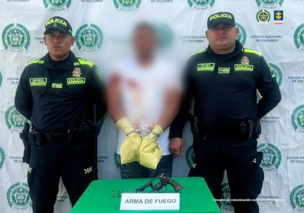 En la imagen aparece una persona de suéter blanco, entre dos uniformados de la Policía. Tiene las manos esposadas y con unas bolsas amarillas que las protegen.
