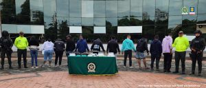 Se observa una fila de 11 personas capturadas, todos de espaldas y a cada lado de la fila dos funcionarios de Policía Nacional. Adelante una mesa con material incautado