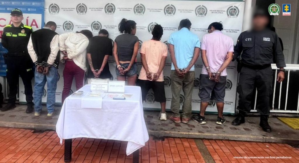 Se observan 7 personas capturadas, de espaldas. A un costado un funcionario del CTI y al otro uno de Policía Nacional que los custodian. Al frente una mesa con material incautado. Atrás un pendón de la Fiscalía General de la Nación y otro de Policía Nacional.