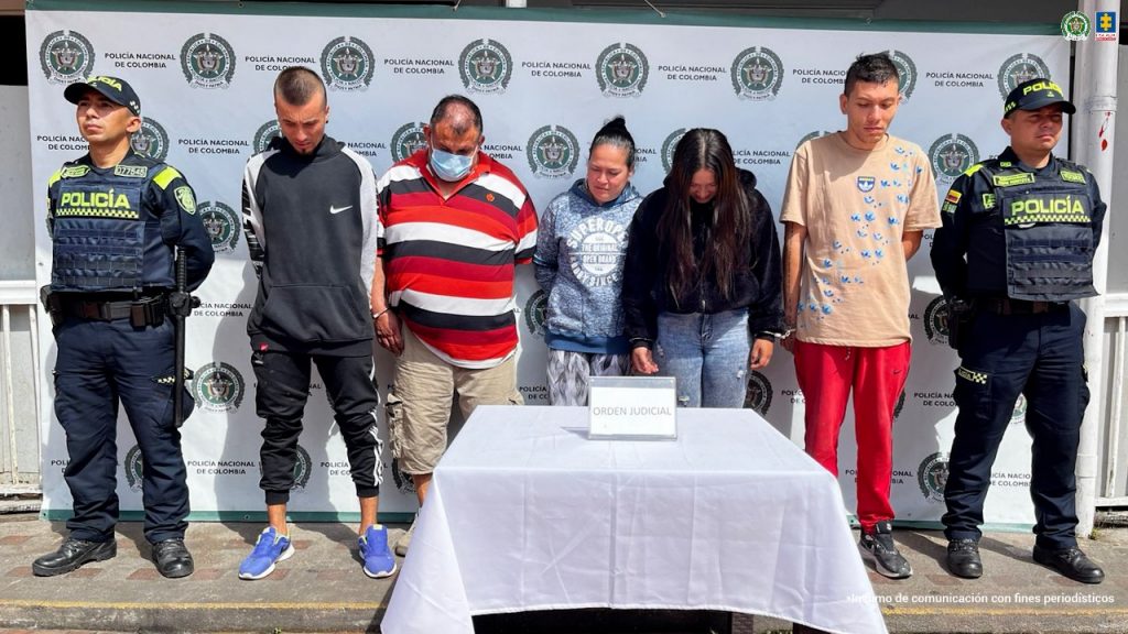 Se observan los cinco capturados de frente, en fila. A cada costado un funcionario de Policía Nacional, atrás, un pendón de Policía Nacional, adelante, una mesa con el cartel ORDEN JUDICIAL