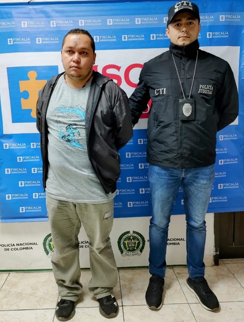 Óscar Arbey Yarce Berrio está en la foto acompañado por un funcionario del CTI de la Fiscalía y en la parte de atrás se ve backing con los logos de la Fiscalía.