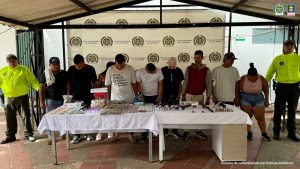 Capturados de frente, custodiados por policías. Detrás banner de la Policía Nacional. Delante una mesa con material incautado en allanamientos.