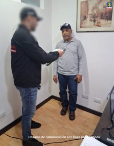 La fotografía está tomada en interiores. Aparece de pie Octavio Hurtado Waitotó. Usa gorra de color negro con las gafas sobre la visera. Viste con camibuso de color gris, pantalón negro y zapatos negros. Frente a él hay un servidor del CTI de la Fiscalía en acción de lectura de un documento que tiene en sus manos.