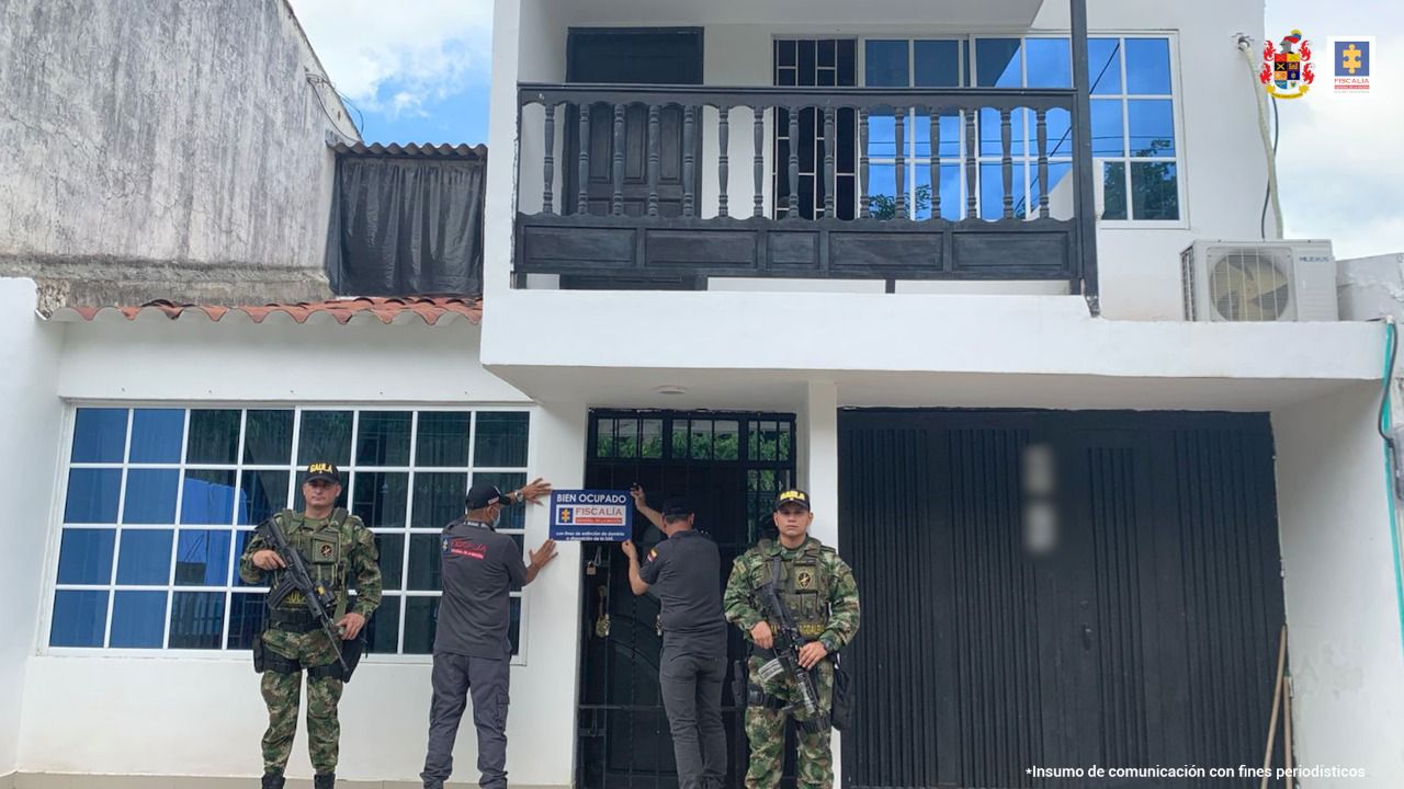 Al fondo es´ta la vivienda afectada con las medidas cautelares. En frente hay dos personas del CTI sosteniendo un cartel que dice Fiscalía y a cada lado hay una persona del ejército. 