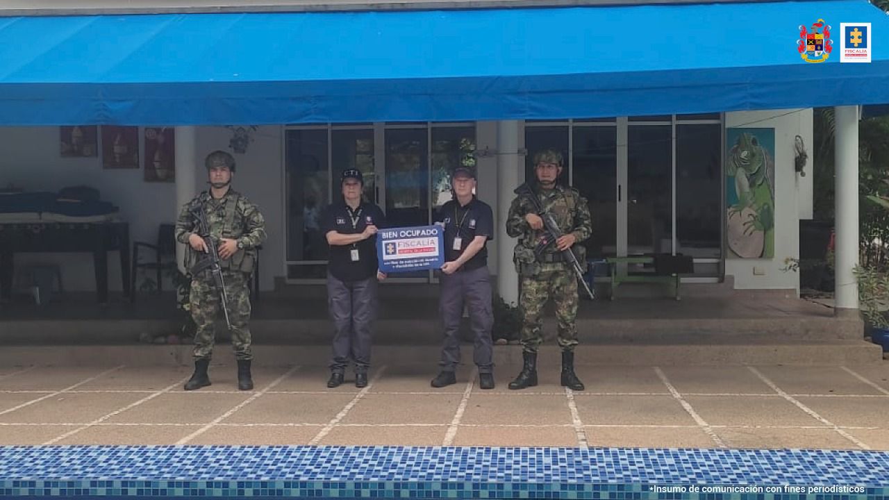 Al fondo es´ta la vivienda afectada con las medidas cautelares. En frente hay dos personas del CTI sosteniendo un cartel que dice Fiscalía y a cada lado hay una persona del ejército. 