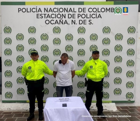 Se visualiza al capturado junto a dos uniformados de la Policía Nacional. Frente a ellos un teléfono celular incautado. Detrás de ellos el banner que identifica a la Policía Nacional.