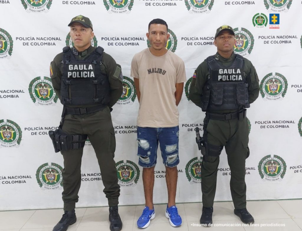 En la imagen aparece una persona con bermuda de jean y camiseta café, entre dos uniformados de la Policía Nacional. 