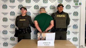 En la fotografía se aprecia de pie, de espaldas a la cámara y esposado a Omar Gil Pérez. Viste camiseta de manga corta de color verde y pantalón azul claro. A sus extremos hay uniformados de la Policía Nacional. Delante de ellos hay una mesa y sobre ella hay un contenedor de cartón y dentro de él se aprecia un arma de fuego. Asimismo, hay un letrero que dice: ‘Armas de fuego’. Detrás de ellos hay un pendón institucional de la Policía.