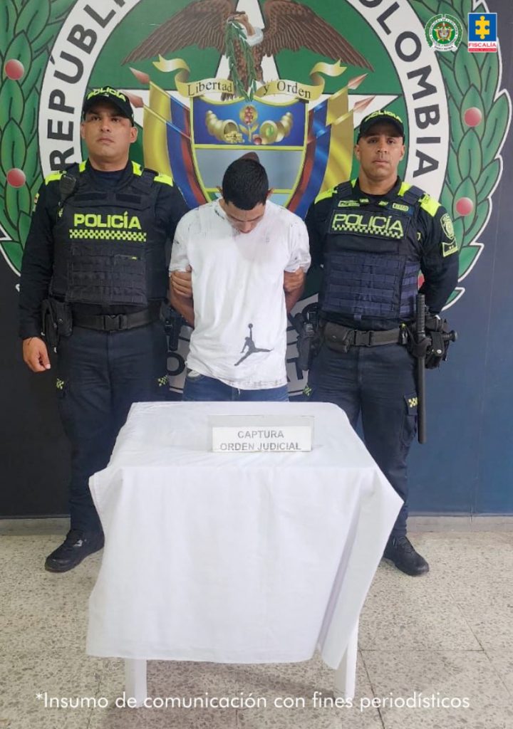En la fotografía se aprecia de pie, los brazos atrás y la cabeza agachada a Omar José Trujillo. Viste con camiseta de color blanco, de manga corta; jean azul. A sus extremos hay uniformados de la Policía Nacional. Delante de ellos hay una mesa con mantel blanco y sobre ella hay un letrero que dice: ‘Captura orden judicial’. Detrás de ellos hay un escudo de la Policía Nacional.