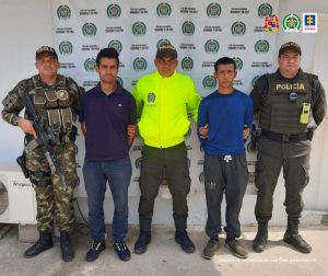 en la imagen se ven dos hombres detenidos bajo custodia de dos integrantes de la Policía Nacional y uno del Ejército. Detrás suyo un backing de la Policía.