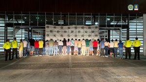 se observa una fila de 27 personas capturadas, de espaldas. A cada costado, dos funcionarios de la Policía Nacional. Atrás un pendón de Policía Nacional.
