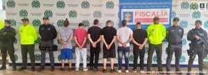 Se observan seis capturados, de espaldas. A cada costado un funcionario del Cuerpo Técnico de Investigación CTI y dos de Policía Nacional. Atrás un pendón de la Fiscalía General de la Nación y uno de Policía Nacional.