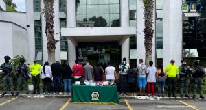 Se observan 12 personas capturadas en fila y todos de espaldas. A cada lado de la fila, tres uniformados de la Policía Nacional. Al frente una mesa con mantel verde y logotipo de la Policía en la cual se ubican elementos incautados en las diligencias de allanamiento .