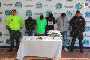 Se observan cuatro hombres detenidos, de frente y en fila. A cada costado de la fila un uniformado de Policía Nacional. Atrás un pendón de Policía Nacional. Al frente una mesa en donde están ubicados los elementos incautados en diligencias de allanamiento.