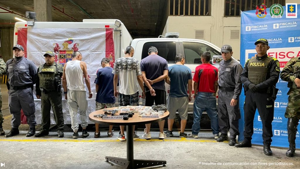 Se observan seis personas capturas, en fila y de espaldas. En casa costado uniformados del CTI y de Ejército Nacional. Al fondo un pensón de Fiscalía General de la Nación y uno de Ejército Nacional, así como una unidad móvil. Al frente una mesa con material incautado en las diligencias de allanamiento
