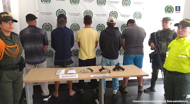 se observan cinco personas detenidas en fila. A un costado un funcionario de la Policía y al otros dos funcionarios de Policía. Al fondo un pendón de Policía Nacional. Al frente una mesa con elementos incautados durante operativos