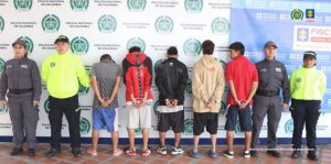 Se observan cinco hombres capturados, en fila, todos de espaldas. A cada costado un funcionario del Cuerpo Técnico de Investigación (CTI) y otro de Policía Nacional. Atrás un pendón de la Fiscalía General de la Nación y uno de la Policía Nacional.
