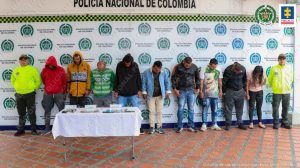 Se observan nueve capturados en fila y a cada extremo un uniformado de la Policía Nacional.  Al fondo un pendón de Policía Nacional.  Al frente una mesa con el material incautado en los allanamientos