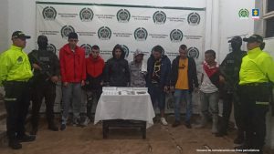 Se observan siete personas capturadas, en fila, de frente.  A cada costados dos funcionarios de Policía Nacional.  Atrás un pendón de Policía Nacional.  Adelante una mesa con material incautado en las diligencias de allanamiento y registro