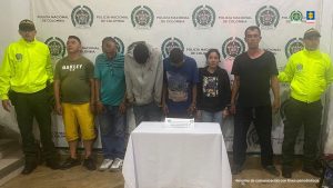 Se observan seis personas capturadas, de frente. A cada costado un funcionario de la Policía Nacional. Atrás un pendón de Policía Nacional.