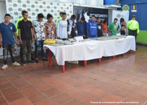 Los imputados se encuentra en custodia por dos uniformados de la Policía Nacional. Detrás se observa un backing de la Policía y se observa la mesa con el material y sustancias de alucinógenos incautados.