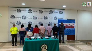 se observan seis capturados en fila, a un costado un funcionario de CTI y al otro uno de Policía nacional. Atrás un pensón de Fiscalía y otro de Policía. Al frente una mesa con material incautado