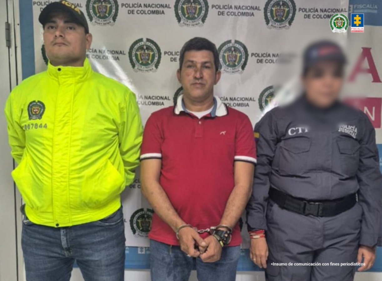 Capturado de frente custodiado por investigador del CTI de la Fiscalía y un policía. Detrás banner de la Fiscalía.