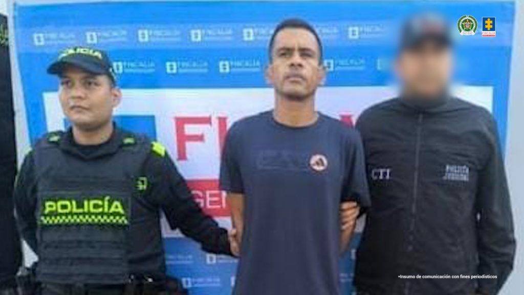 Se visualiza al capturado junto a un uniformado de la Policía Nacional y un investigador del CTI. Detrás el banner que identifica a la Fiscalía General de la Nación.
