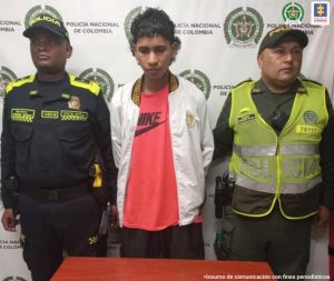 Dos efectivos de la Policía Nacional custodian al procesado quien está esposado. De fondo el backing de la Policía.