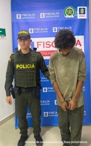 Se observa al capturado junto a uniformado de la Policía Nacional. En la parte posterior se ubica el banner que identifica a la Fiscalía General de la Nación.