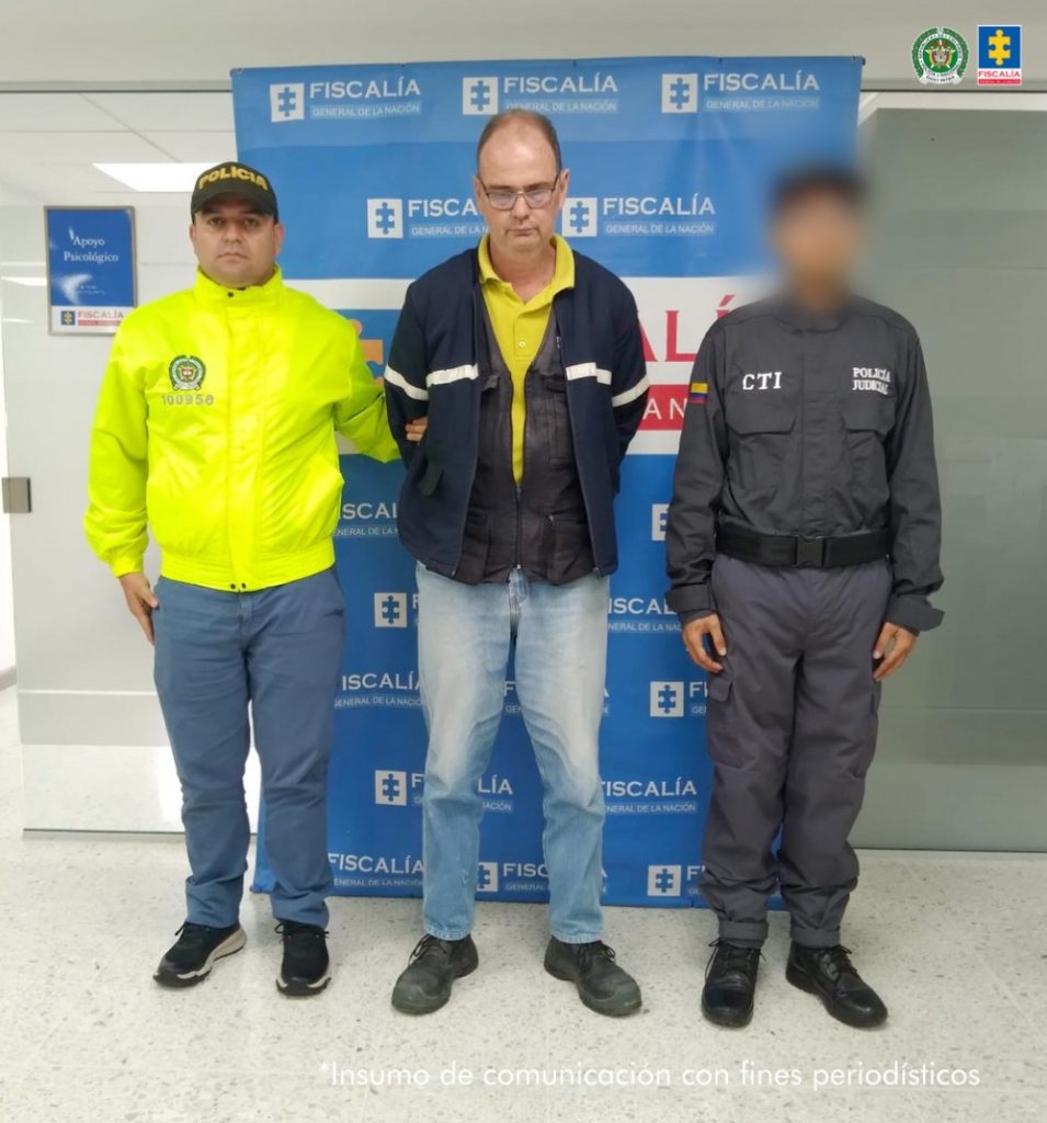 en la fotografía se puede observar al detenido en medio de un agente del CTI y un funcionario de la policia, detras de ellos hay un baking de la Fiscalia