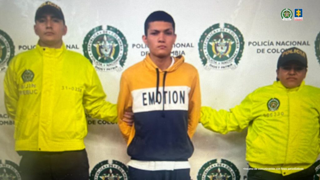 En la fotografía se puede ver al capturado en medio de dos funcionarios de la policia, detras de ellos hay un baking de la policia