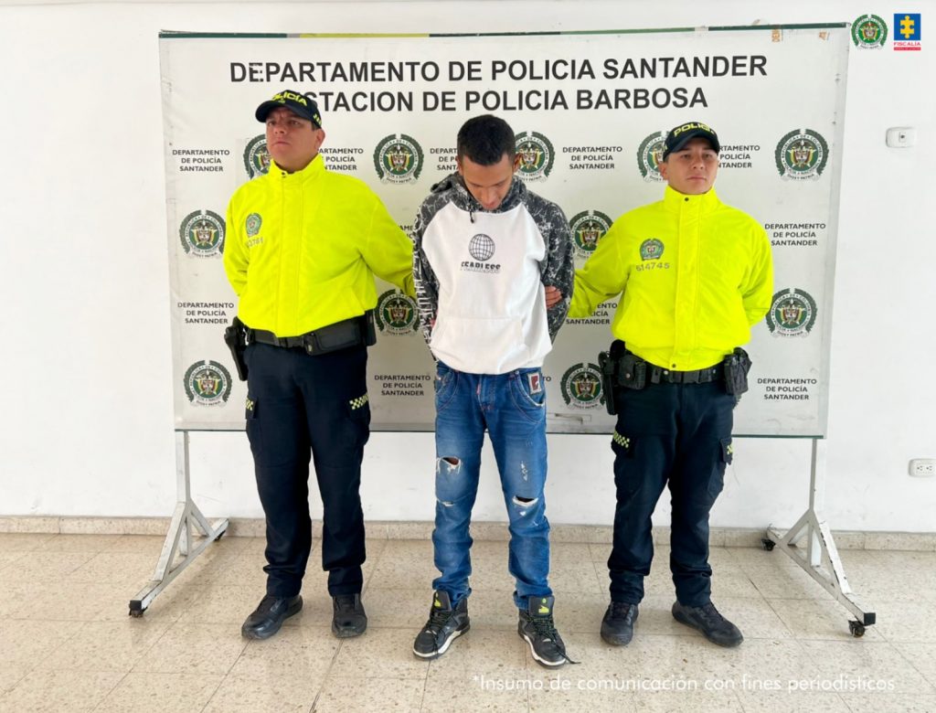 "Un hombre esposado es custodiado por dos policías uniformados frente a un cartel institucional que dice 'Departamento de Policía Santander - Estación de Policía Barbosa'. Los policías visten chaquetas amarillas fosforescentes. Al fondo se observan logotipos oficiales de la Policía Nacional y la Fiscalía General de la Nación."