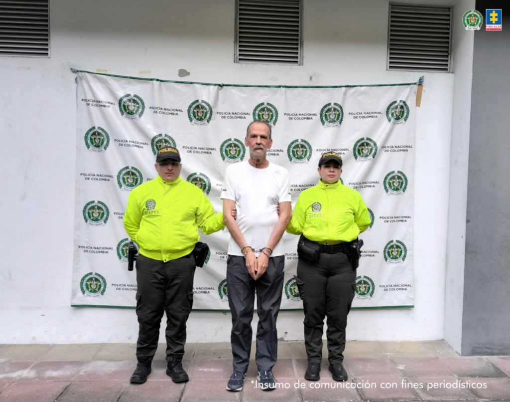 En la fotografia se ve al detenido en medio de dos policias, atras de ellos un baking de la policia