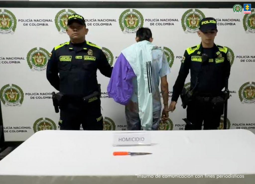 En la fotografía se puede observar al capturado de espaldas en medio de dos policias que lo custodian. Delante de ellos hay una mesa en donde se puede ver el arma cortopunzante con el que cometio el delito y detras de todo un baking de la POlicia