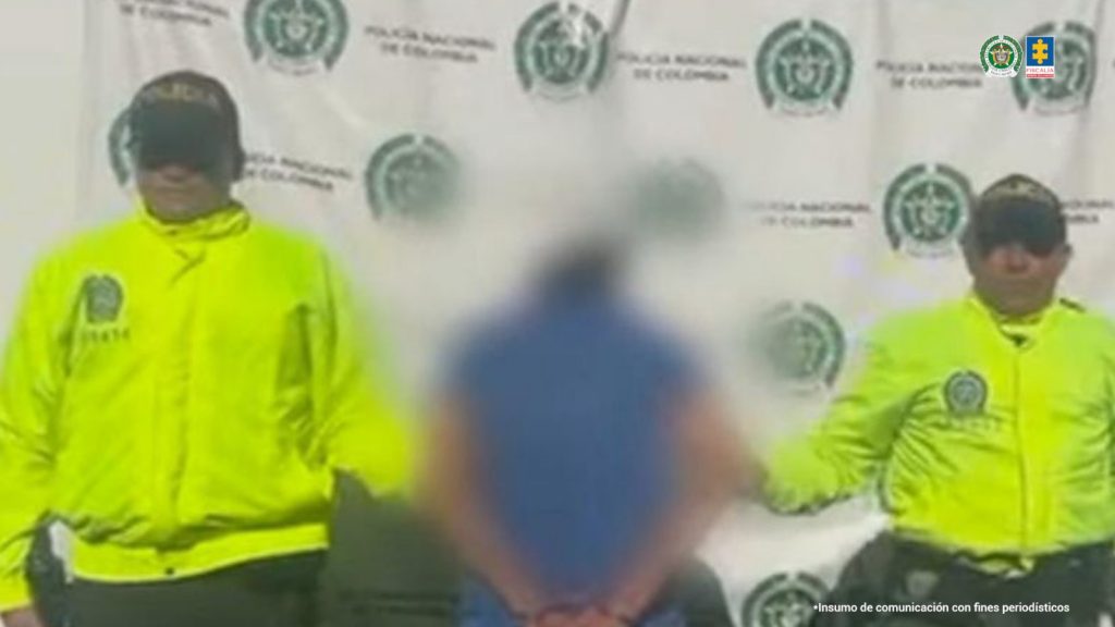 Fotografia en donde se ve al capturado de espaldas difuminado, en medio de dos agentes de policia, detras de ellos un baking de la policia