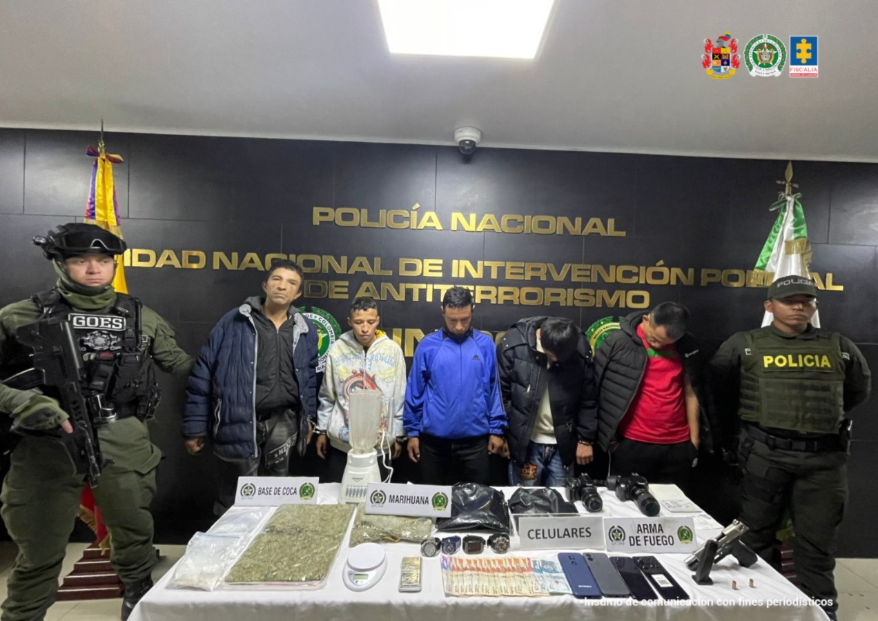 Cinco hombres esposados están de pie en fila, custodiados por dos policías uniformados y un integrante del GOES armado. Frente a ellos, sobre una mesa blanca, se exhiben evidencias incautadas: bolsas con marihuana, base de coca, una licuadora, una báscula, varios celulares, cámaras fotográficas, dinero en efectivo y un arma de fuego. Al fondo se lee un letrero de la Policía Nacional y de la Unidad Nacional de Intervención Policial de Antiterrorismo.