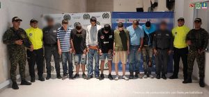 En la foto se observa a un grupo de hombres custodiados por las autoridades.En el centro hay ocho hombres con la cabeza agachada y las manos esposadas al frente, lo que sugiere que se trata de personas detenidas. A cada lado de ellos se ubican integrantes de la Policía Nacional de Colombia, el CTI de la Fiscalía y militares, algunos vestidos con uniformes camuflados y portando armas, otros con chaquetas amarillas de alta visibilidad y uniformes oscuros. El fondo está compuesto por pendones institucionales de la Policía Nacional y la Fiscalía General de la Nación, lo que indica que es una presentación oficial de capturas. La escena corresponde a un procedimiento judicial en el que se exhibe a varios detenidos junto con la fuerza pública.