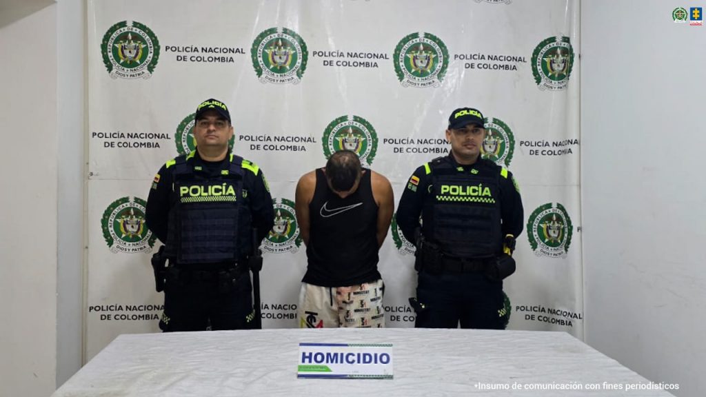 En la fotografia se puede ver al detenido en cusodia de dos funcionarios de la policia, detras de ellos de se ve un pendon de la Policia. y en frente de ellos hay un mesa con un letrero en el que se puede leer "homicidio"