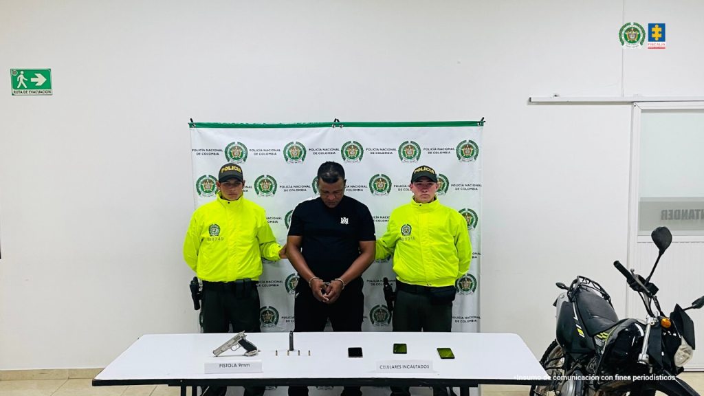 en la fotografia se puede ver al detenido cusodiados por dos funcionarios de la policia, en frente de ellos hay una mesa con elementos incautados y detras de todos ellos hay un pedon de la policia. al lado derecho de la foto se puede ver una moto