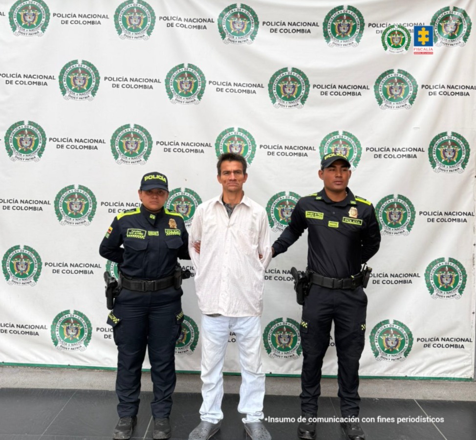 en la fotografia se puede ver al detenido vestido de blanco en medio de dos policias, atras de ellos se ve un pendon de la Policía