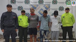 Se observan tres capturados al centro. A un costado de la fila un funcionario de la policía y otro del CTI. Al otro lado un funcionario de Policía. Al fondo un pendón de Fiscalía General de la Nación y uno de Policía Nacional