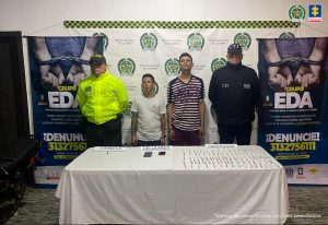 En la fotografía se aprecia de pie a Pablo Andrés Sánchez Menjura, alias ‘Pablito’ y Rubén Darío Martínez, esposados entre sí. Uno, viste camiseta blanca de manga corta. El otro viste camiseta de manga corta con rayas oscuras y blancas, y un estampado en color rojo. Al lado derecho de ellos hay un uniformado de la Policía Nacional. Al lado izquierdo de él hay un servidor del CTI de la Fiscalía. Delante de ellos hay una mesa con mantel blanco y sobre ella se aprecian dos teléfonos celulares, varios cigarrillos y una vainilla. Asimismo, hay tres letreros que dicen: ‘Vainilla’, ‘Celulares’ y ‘Marihuana’. En la parte posterior hay un pendón institucional de la Policía y dos que hacen referencia al Grupo EDA, con un número telefónico para denunciar y logos de la Fiscalía, Policía y Medicina Legal.