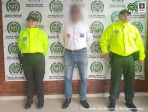 En la imagen el investigado esta de frente, esposado con las manos atrás y custodiado por dos uniformados de la Policía Nacional.