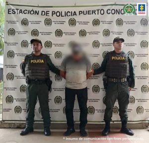 capturado de frente manos atrás esposado, custodiado por dos policías. Detrás banner de la Policía Nacional.