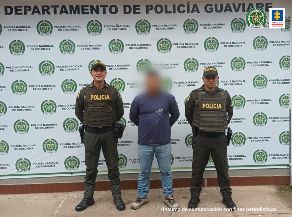 capturado de frente esposado manos atrás, custodiado por dos uniformados de la Policía. Detrás banner de la Policía Nacional.